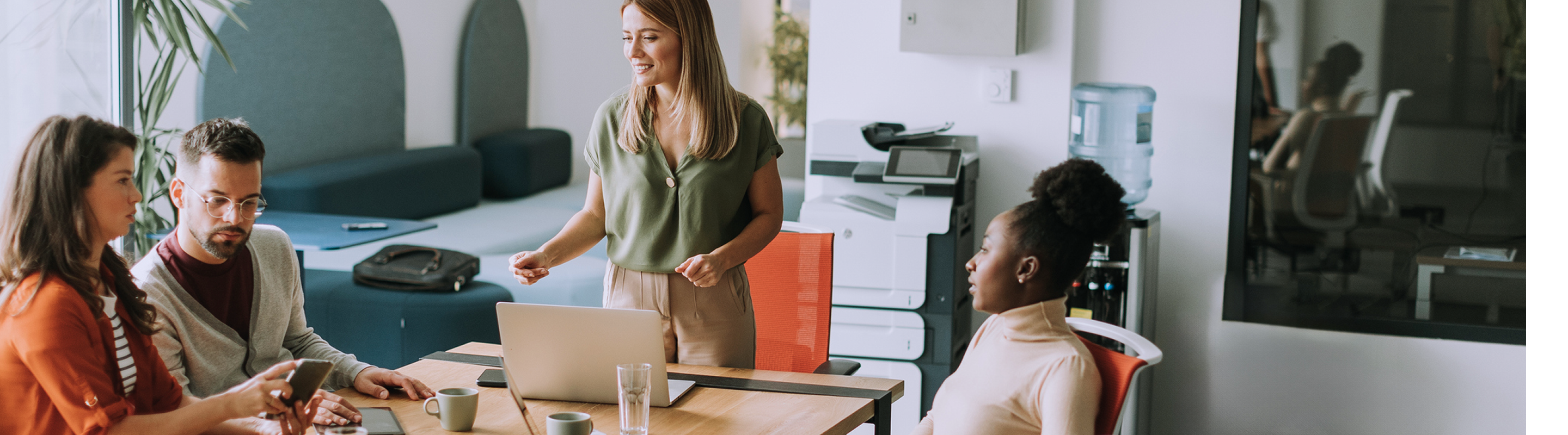 A group of four people engaged in a meeting around a table with laptops and coffee in a modern office setting.